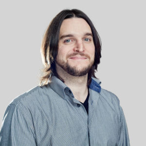 Portrait of a smiling man with shoulder-length hair and a beard in a casual shirt.