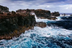 Waves crashing against rugged coastal rocks under a cloudy sky.