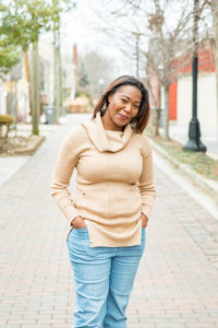 Woman in beige sweater standing on a city sidewalk with hands in pockets.
