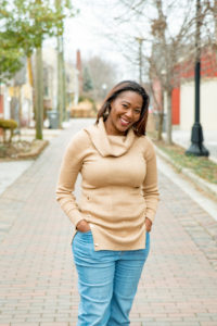 Smiling woman in a beige sweater stands confidently on a city sidewalk with hands in pockets.