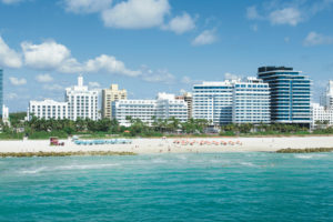 Sunny beach with turquoise water and white buildings in the background.