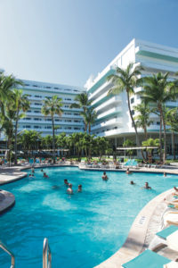 People swimming in a large resort pool surrounded by palm trees and modern buildings.