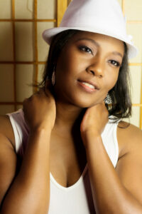 Close-up portrait of a woman gently touching her neck, wearing a white hat and earrings.